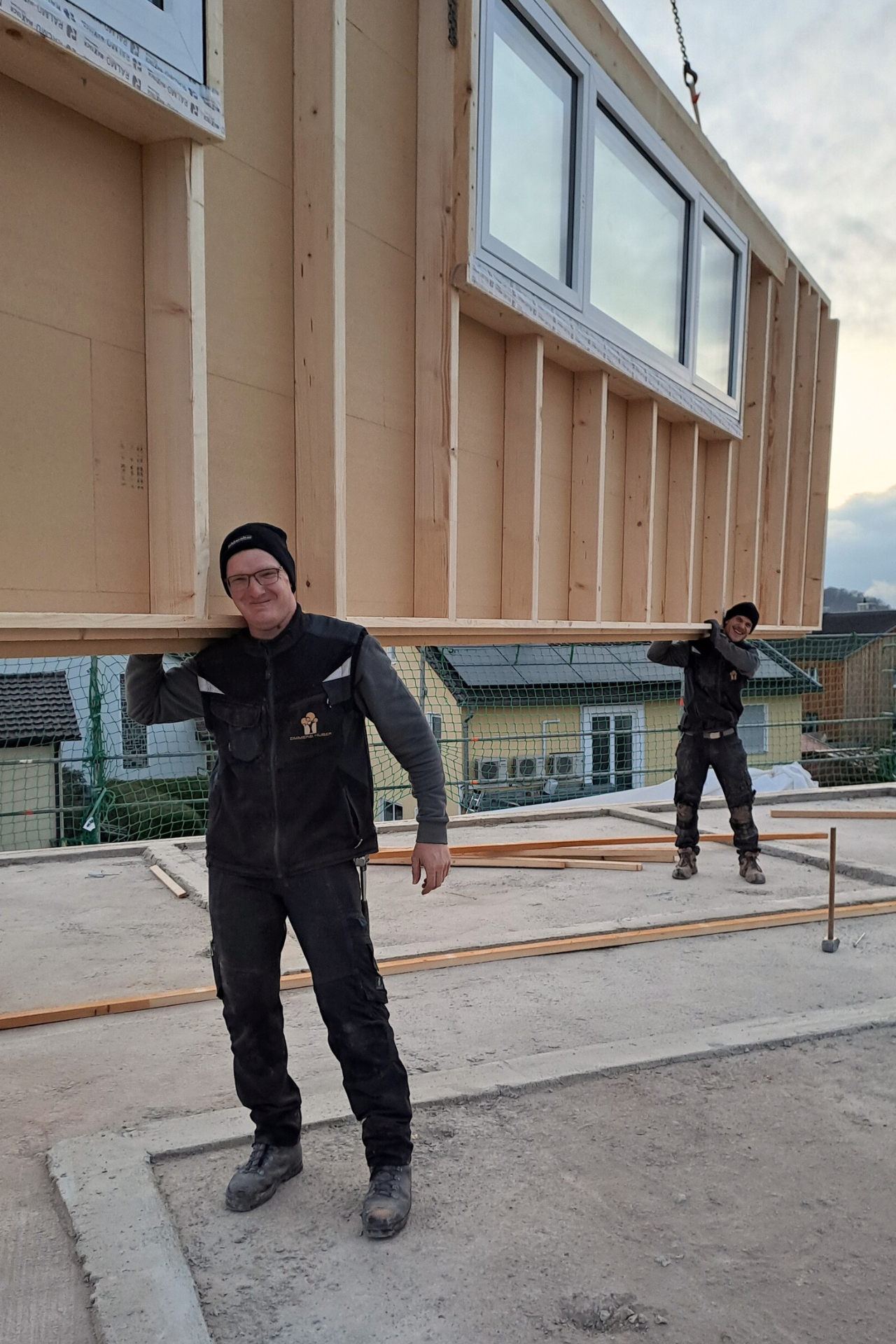 Zwei Mitarbeiter der Zimmerei Huber mit einer am Kran hängenden Hauswand auf den Schultern, der Kran ist außerhalb des Fotos, so dass es aussieht als würden sie die Wand tragen.